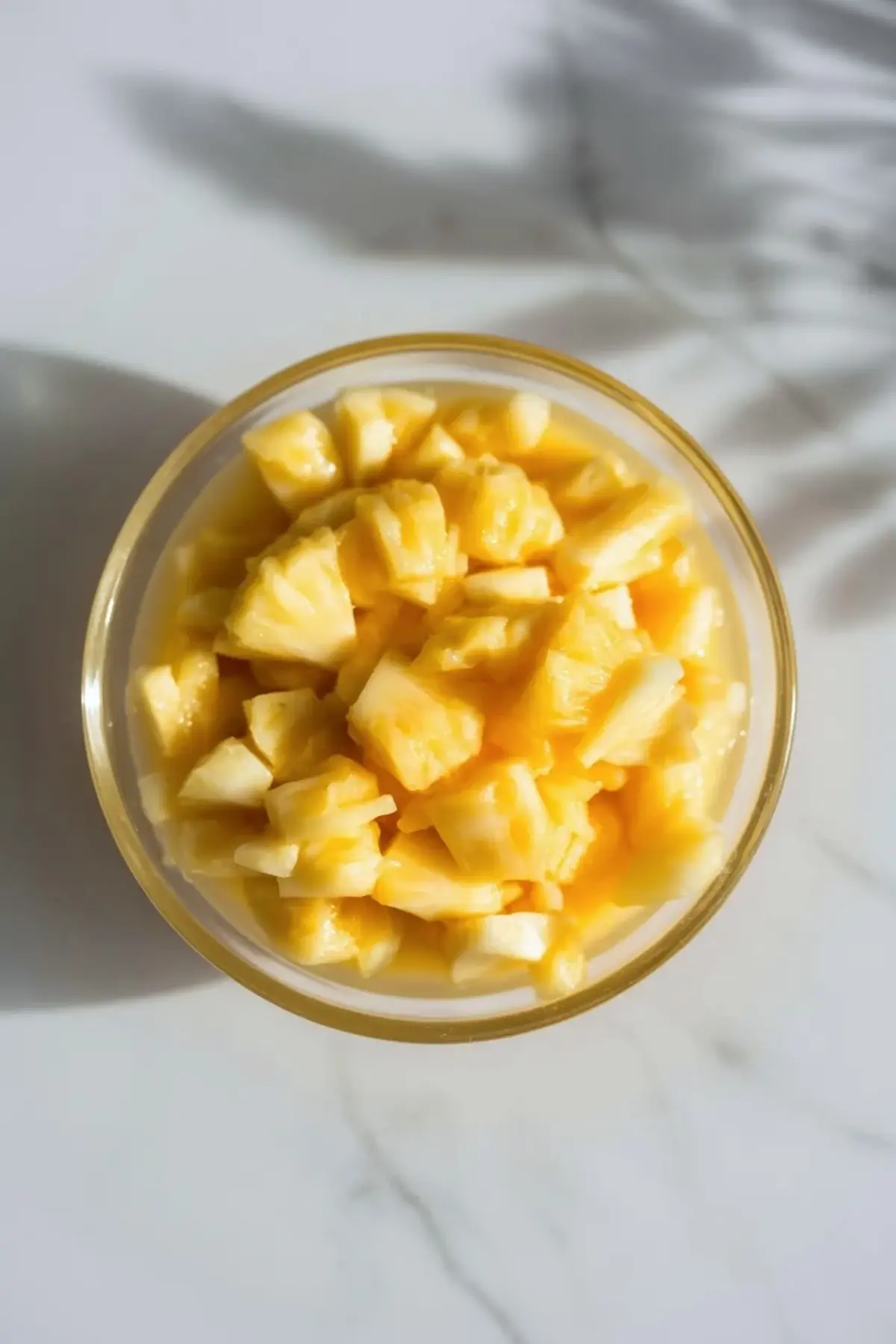 Bowl of pineapple pieces in thick yellow sauce for piña colada dump cake filling on a white marble surface.