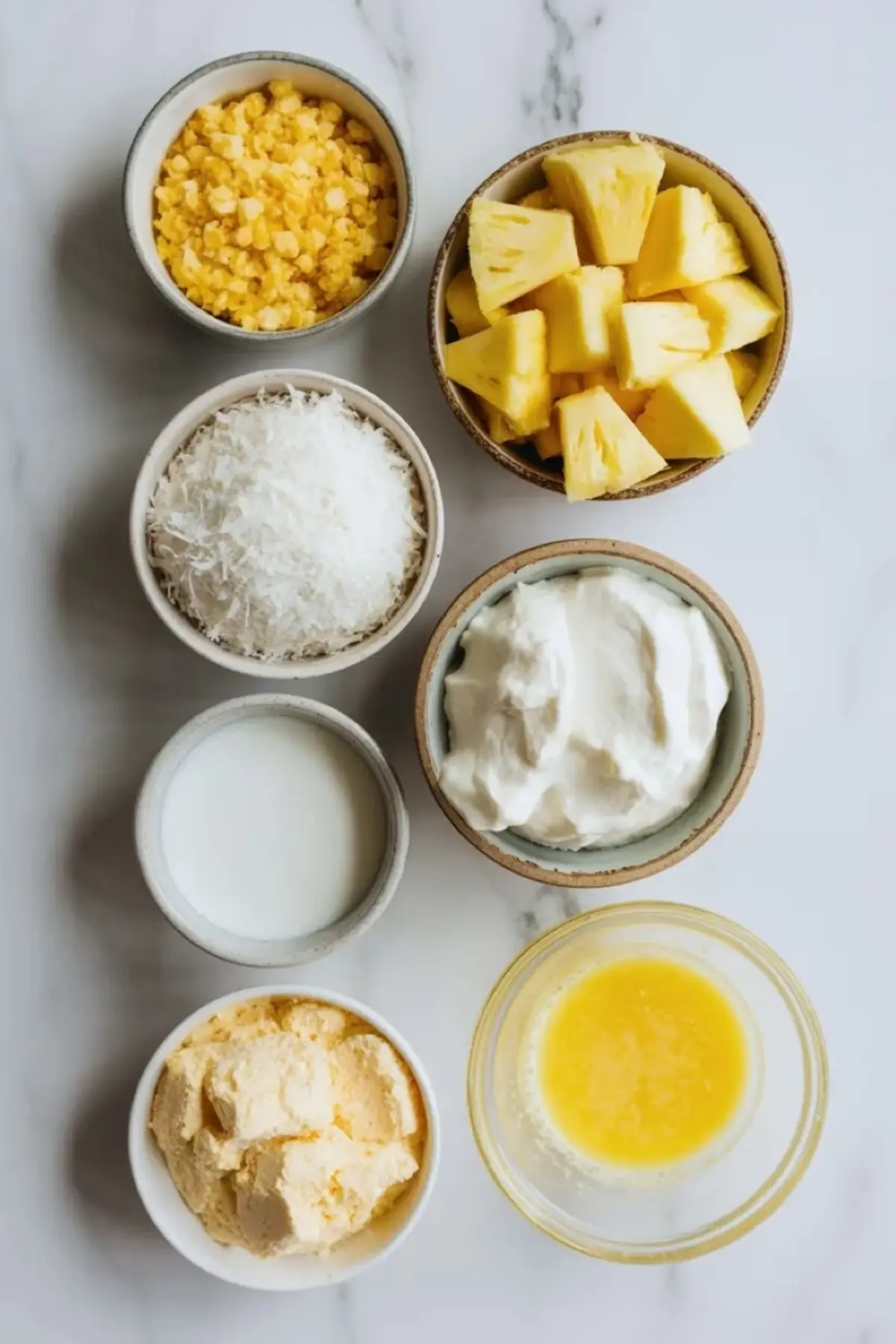 Piña colada dump cake ingredients arranged in bowls with pineapple chunks, shredded coconut, cream, cake mix, and melted butter on white marble.