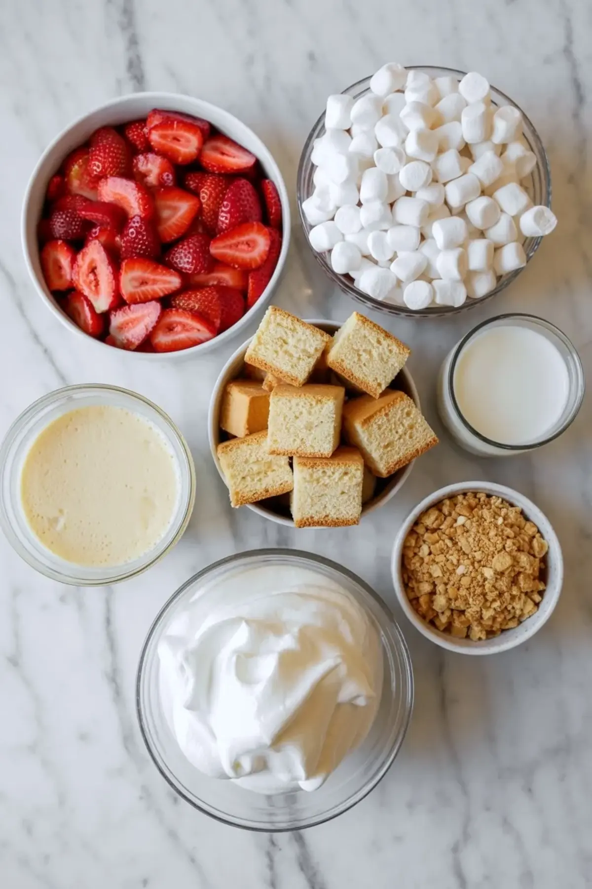Ingredients for strawberry shortcake fluff salad are arranged on white marble with strawberries, mini marshmallows, cake cubes, whipped topping, pudding base, milk, and crushed cookie crumbs.