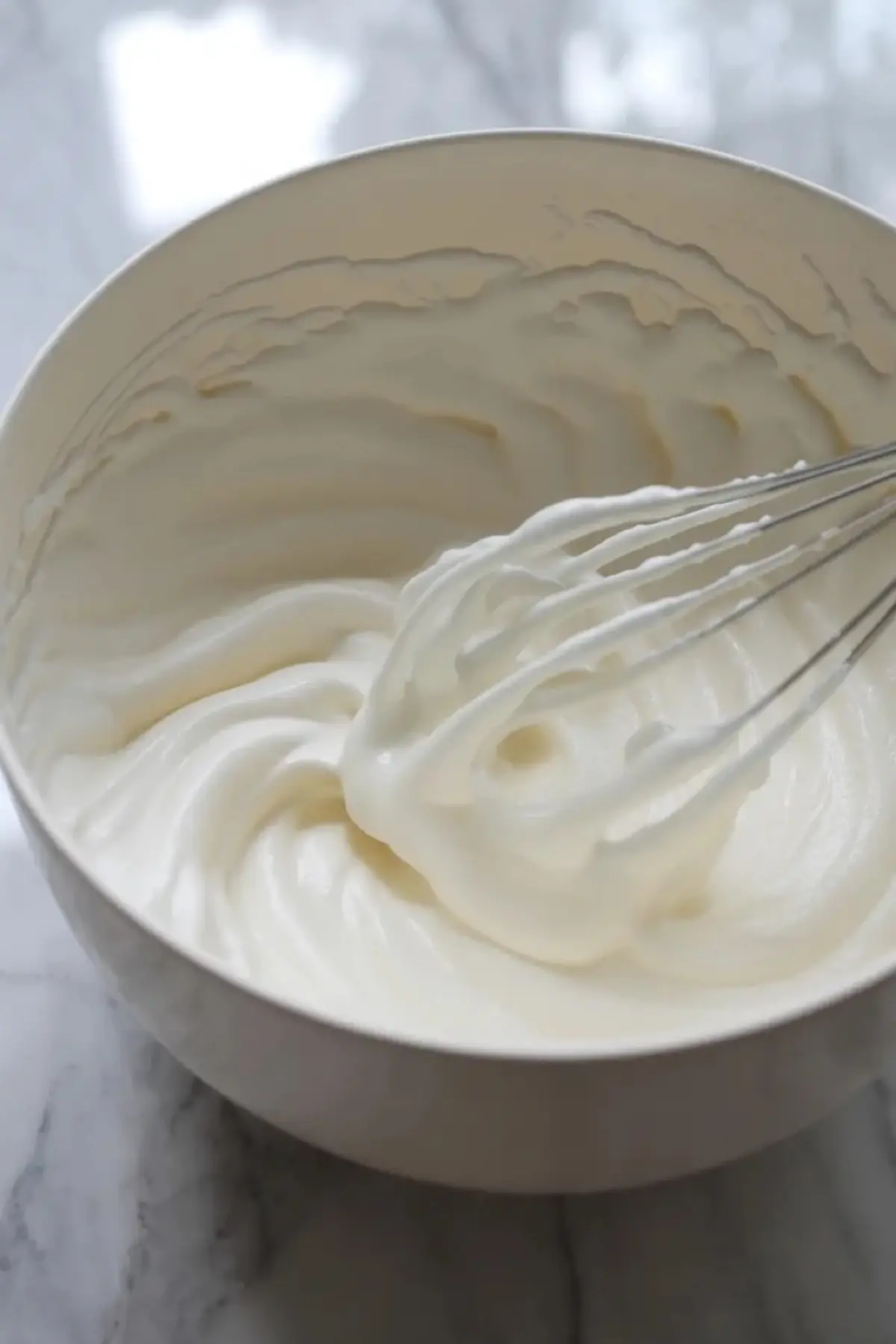 Whipped mascarpone mixture for strawberry tiramisu in a white bowl with a whisk on a marble surface.