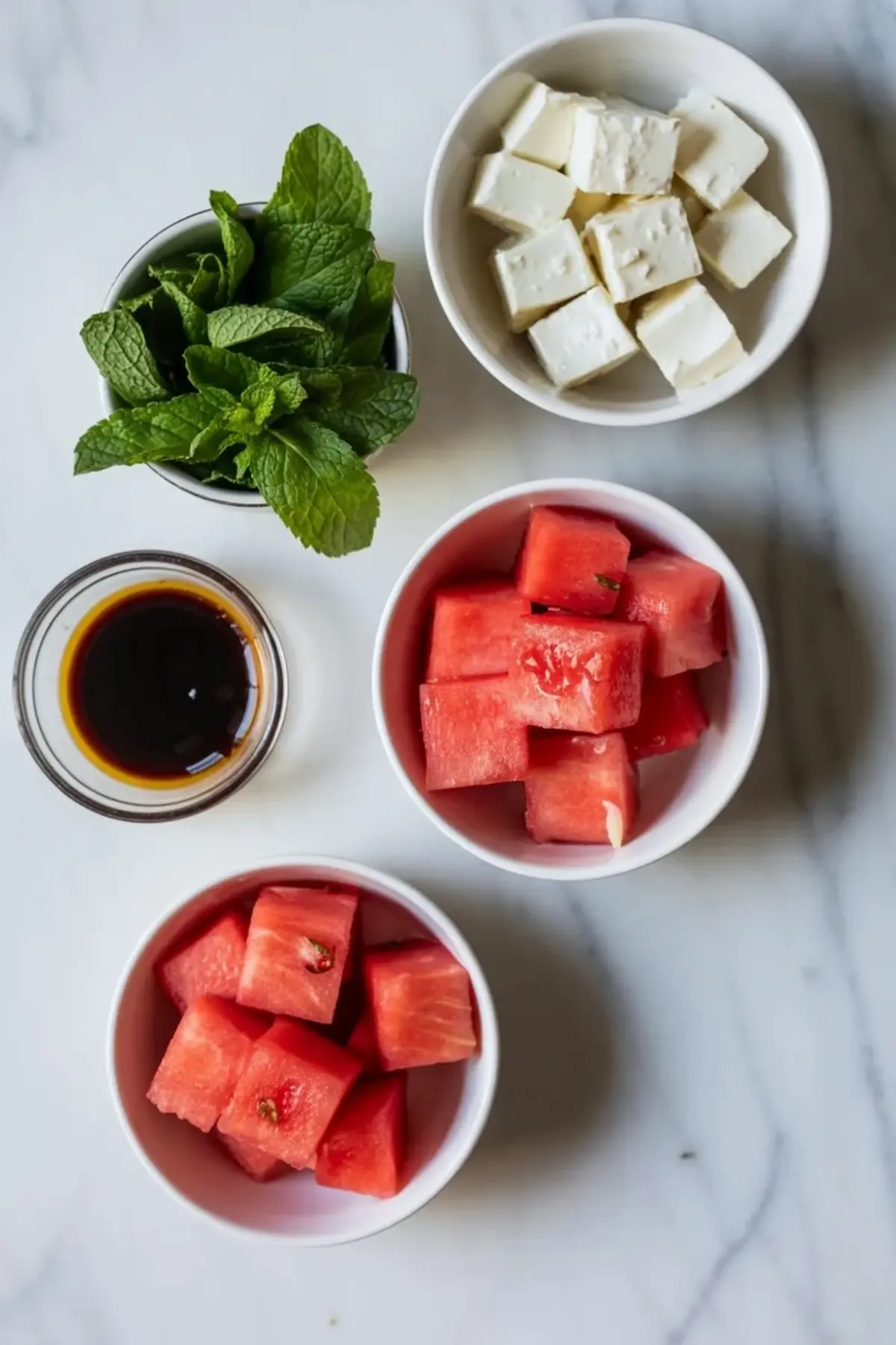 Ingredients for watermelon feta mint skewers arranged on white marble with watermelon cubes, feta cubes, mint, and balsamic glaze.