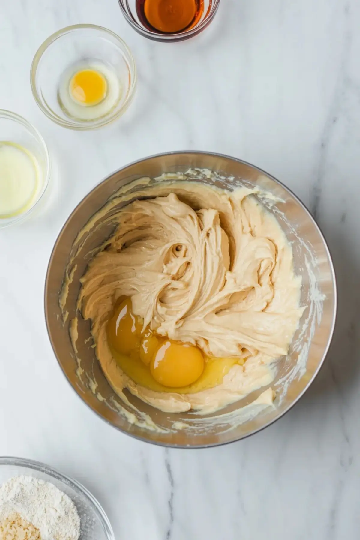 Mixing bowl for almond paste cookies with almond paste mixture and egg being added before the dough is fully combined, surrounded by flour and extracts on white marble.