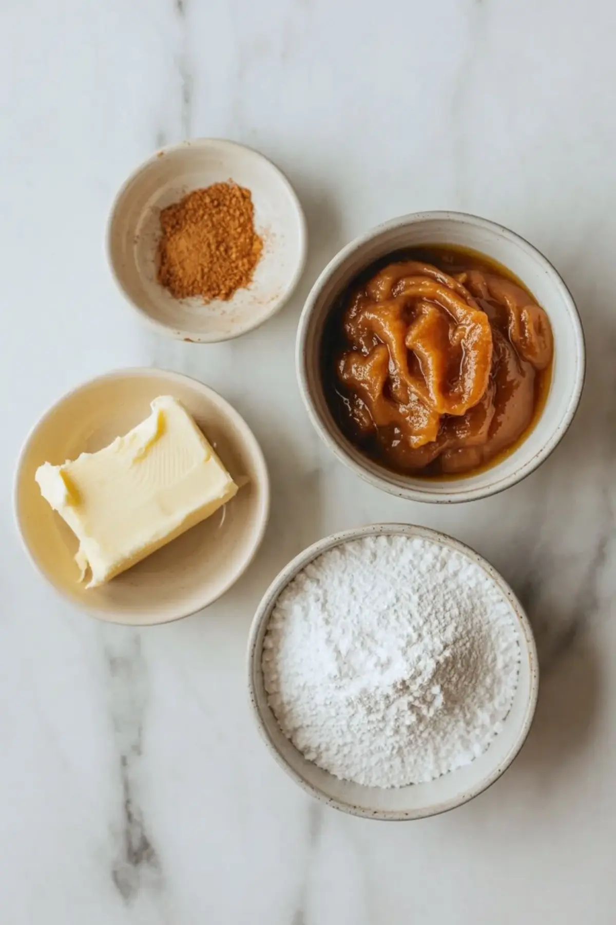 Ingredients for apple butter frosting arranged overhead with butter, powdered sugar, apple butter, and cinnamon on a light surface.