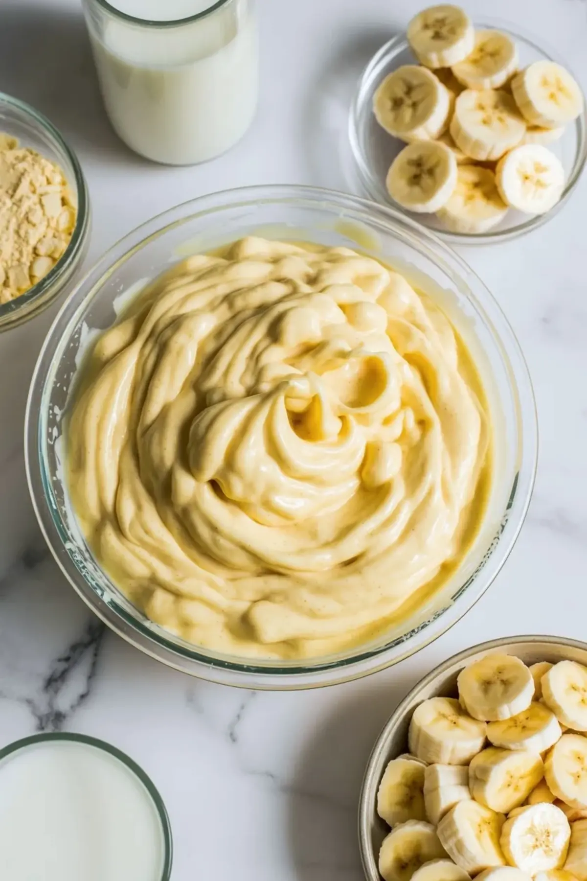 Creamy banana filling for banana cream pie cupcakes in a bowl with sliced banana and milk nearby.