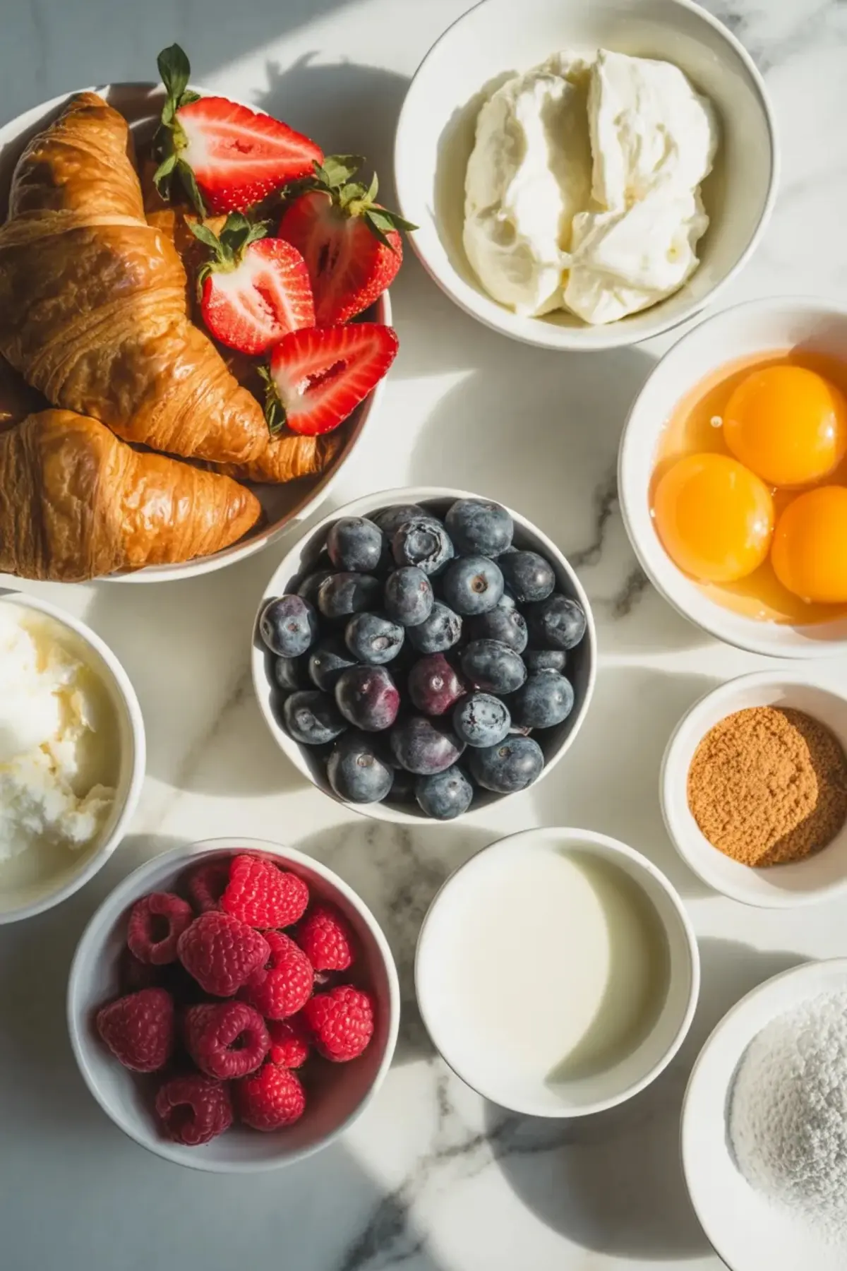 Ingredients for berry croissant breakfast bake with croissants, strawberries, blueberries, raspberries, cream cheese, eggs, milk, and sugar arranged on a white surface.