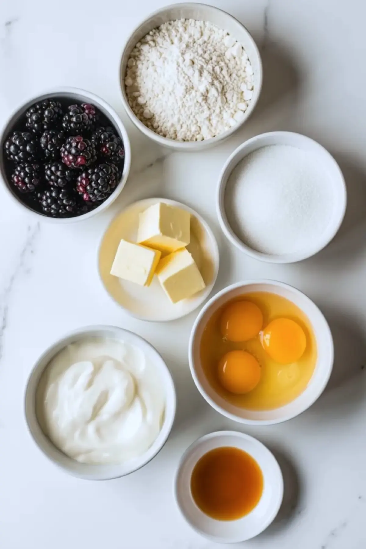 Ingredients for blackberry cobbler bars are arranged on white marble with blackberries, flour, butter, eggs, sour cream, sugar, and vanilla.