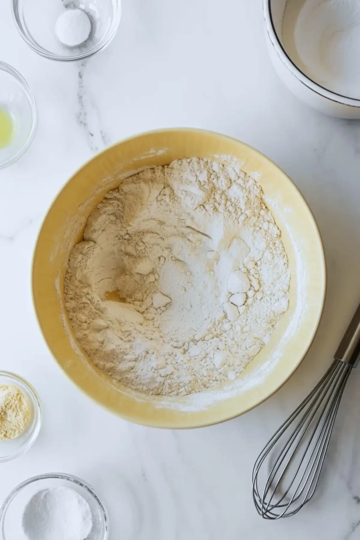 Dry ingredients for chamomile cake in a yellow mixing bowl with flour, leaveners, and nearby prep bowls on a white work surface.