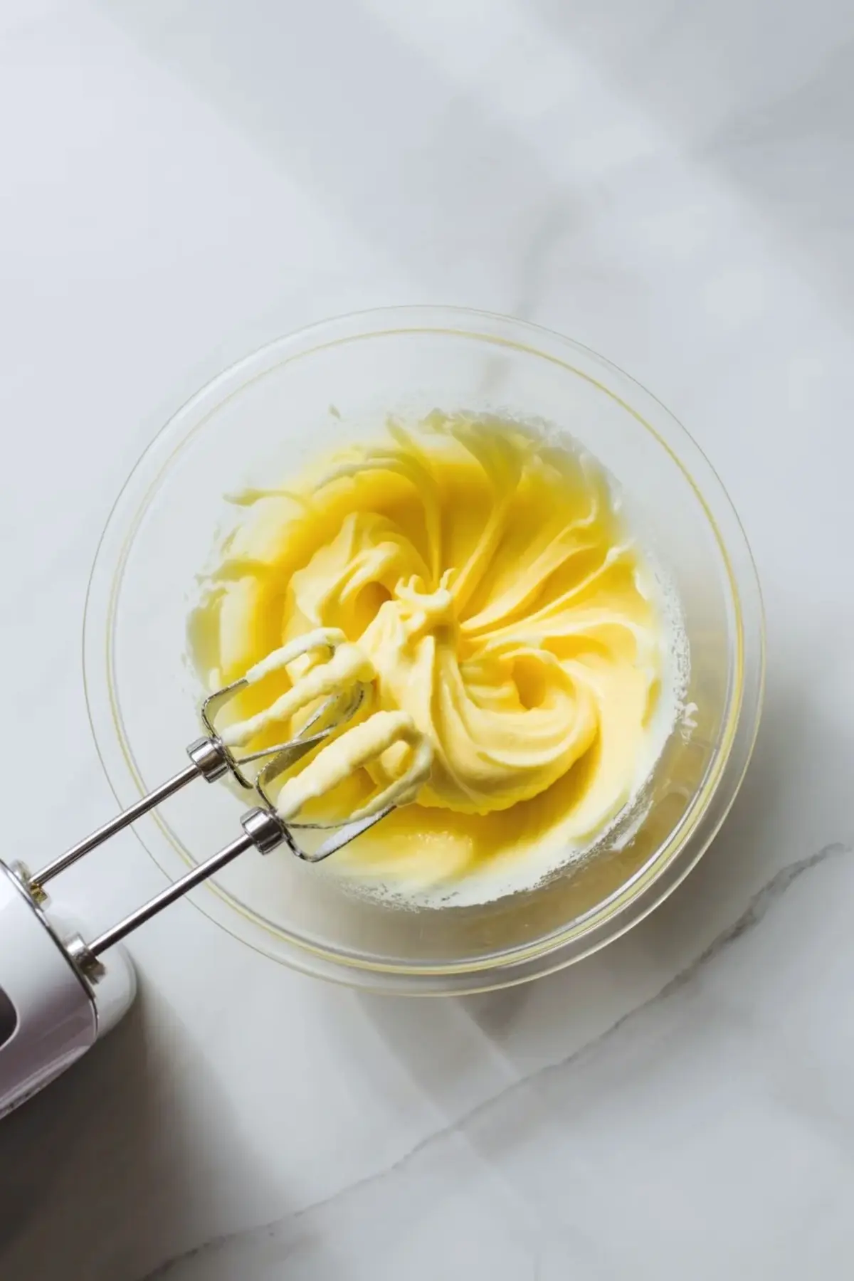 Softened butter being whipped in a clear glass bowl with mixer beaters to start cinnamon buttercream frosting.