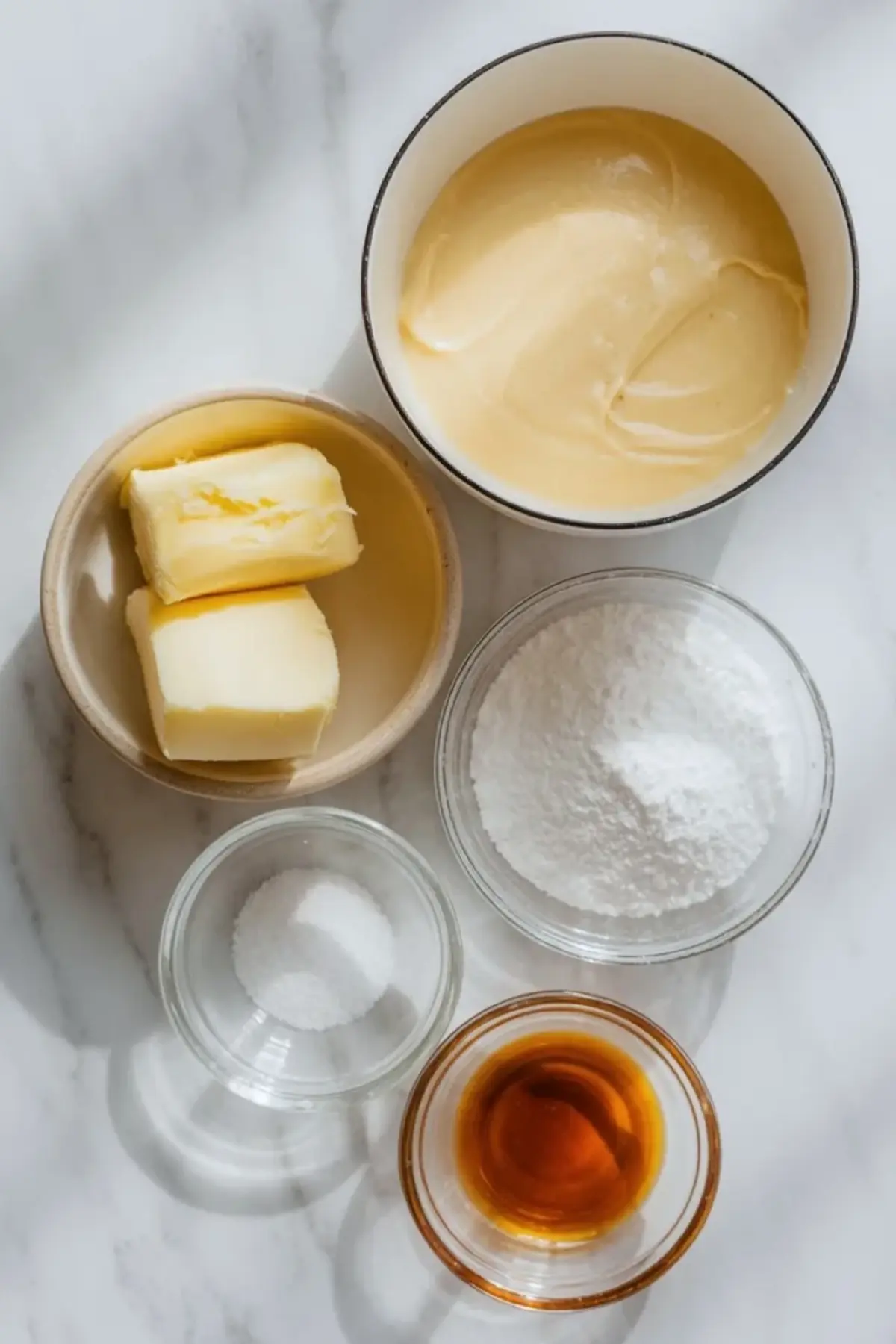 Ingredients for condensed milk frosting arranged overhead with butter, sweetened condensed milk, powdered sugar, salt, and vanilla on a white marble surface.