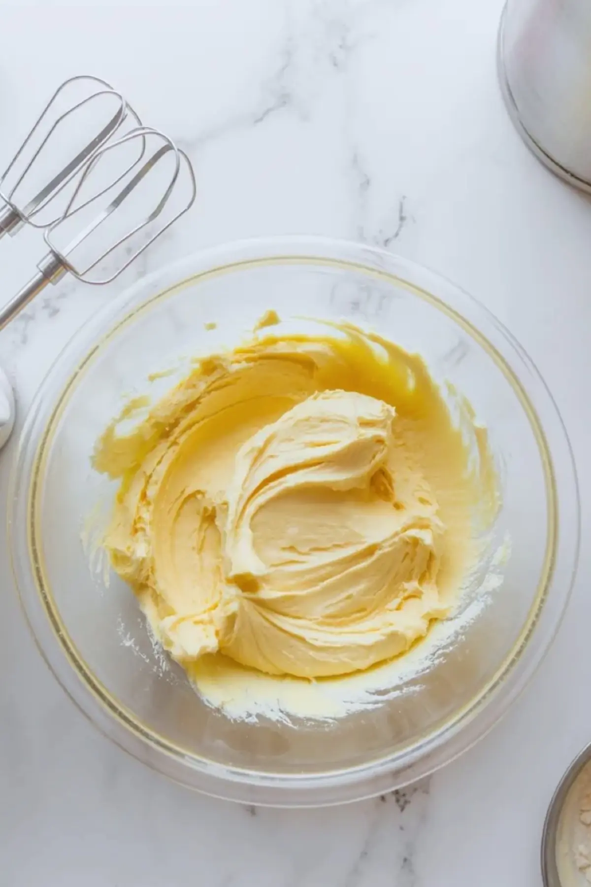 Creamed butter for cookie butter frosting sits in a glass mixing bowl with mixer beaters nearby on a white marble surface.