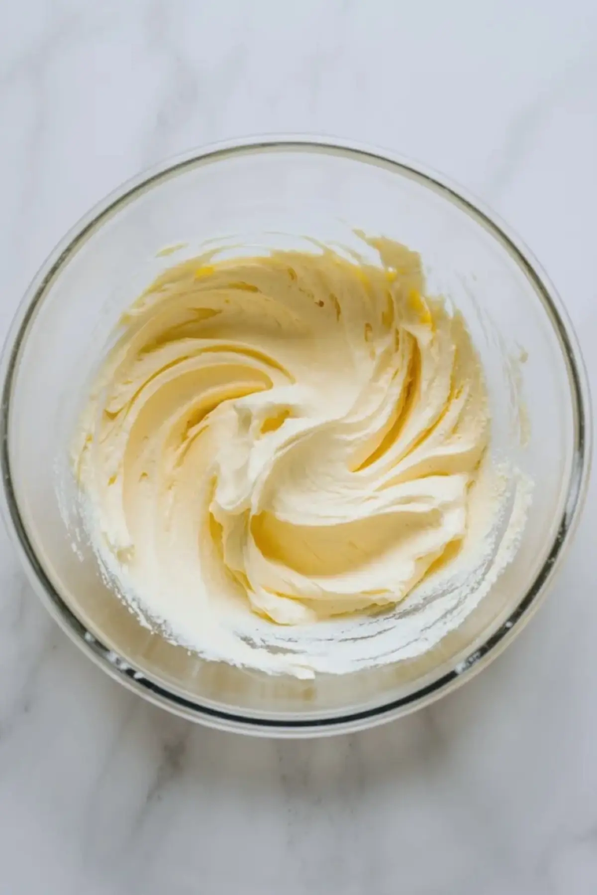 Softened butter whipped in a glass mixing bowl to start dark chocolate buttercream frosting on white marble.