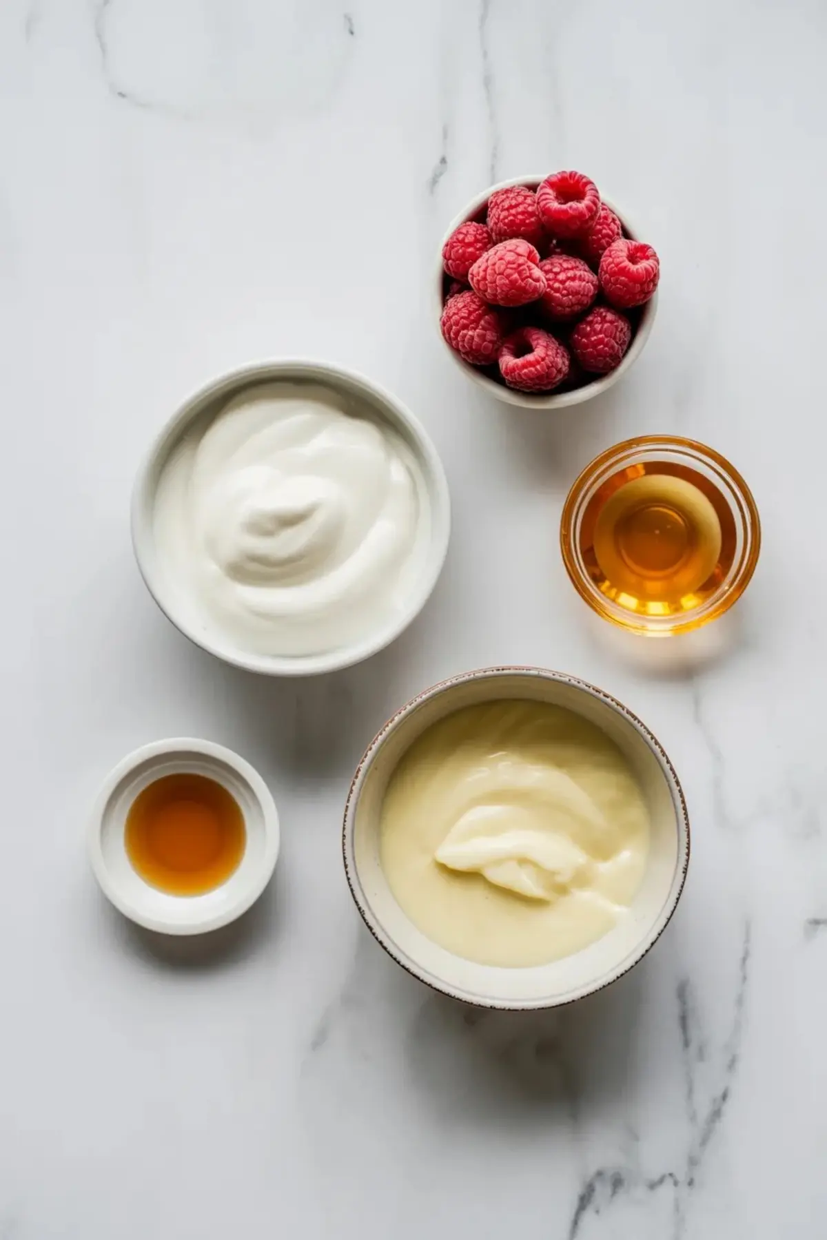 Ingredients for frozen yogurt raspberry bites on white marble with Greek yogurt, raspberries, honey, vanilla, and small bowls for mixing.