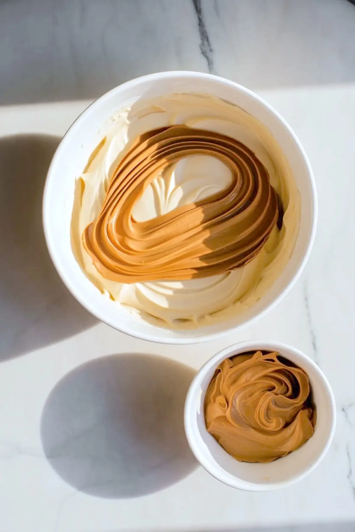 Hazelnut praline paste swirled into the frosting base in a glass bowl for hazelnut praline cream frosting with extra praline nearby.