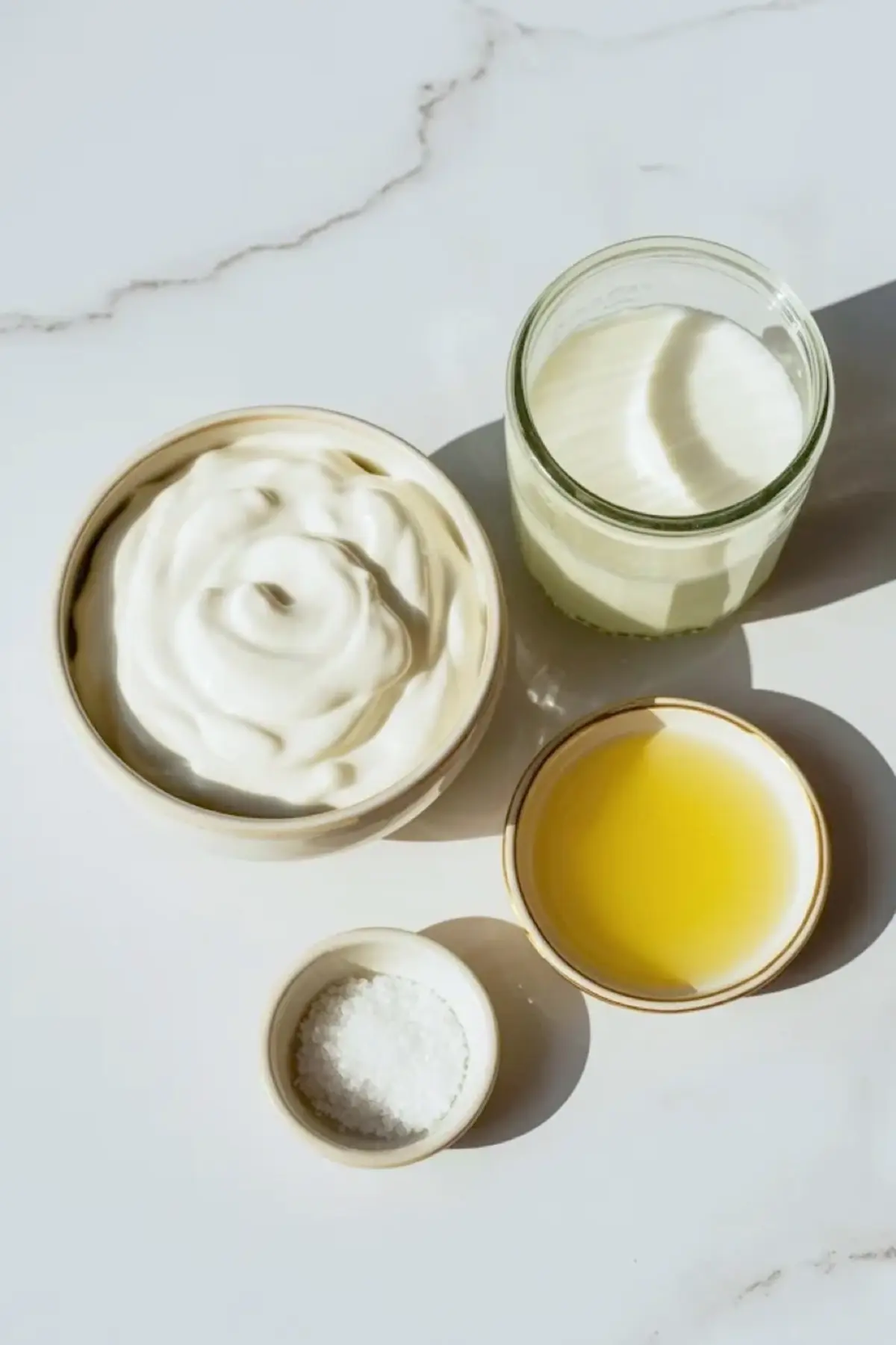 Ingredients for homemade sour cream with heavy cream, cultured buttermilk, lemon juice, and salt arranged on a white surface beside finished sour cream.