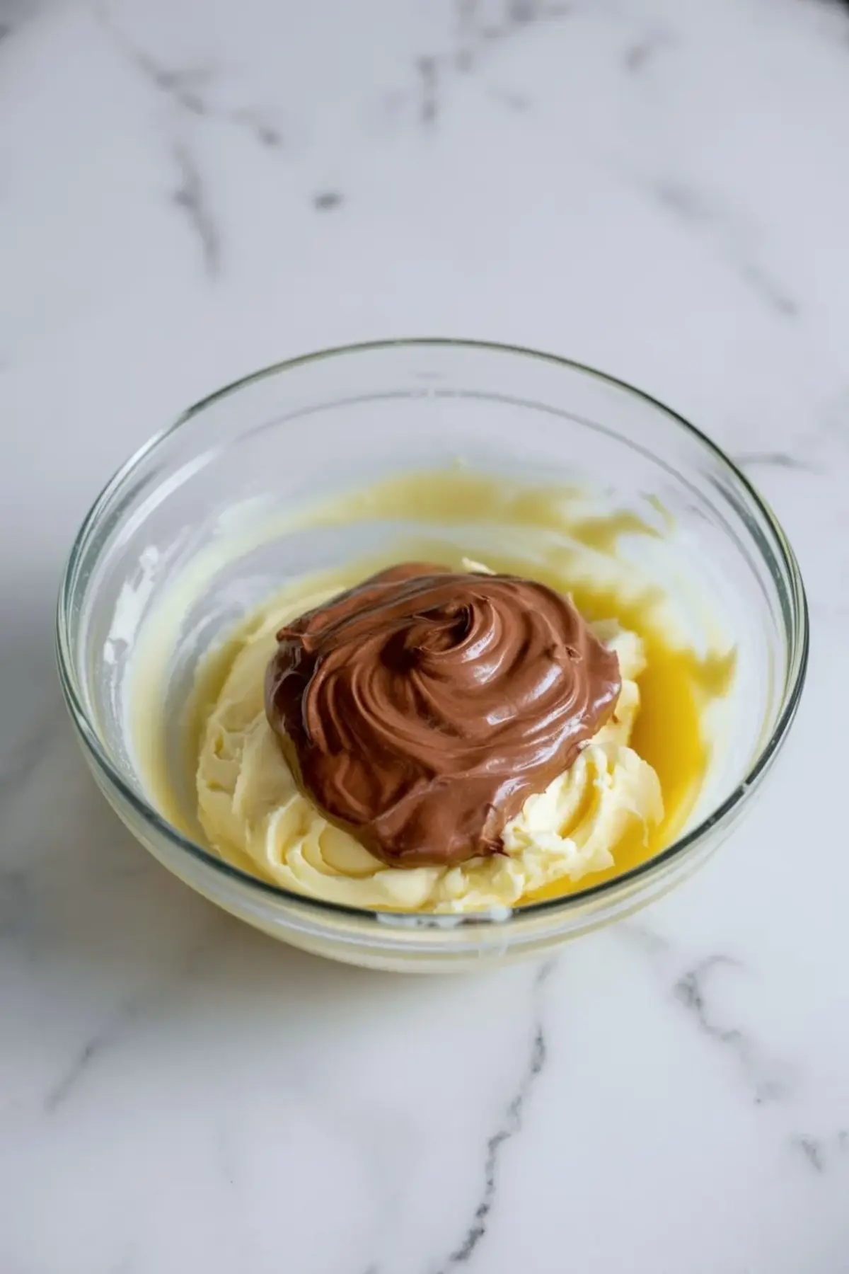 Nutella added to whipped butter in a glass mixing bowl for Nutella buttercream frosting before the powdered sugar and cream are mixed in.