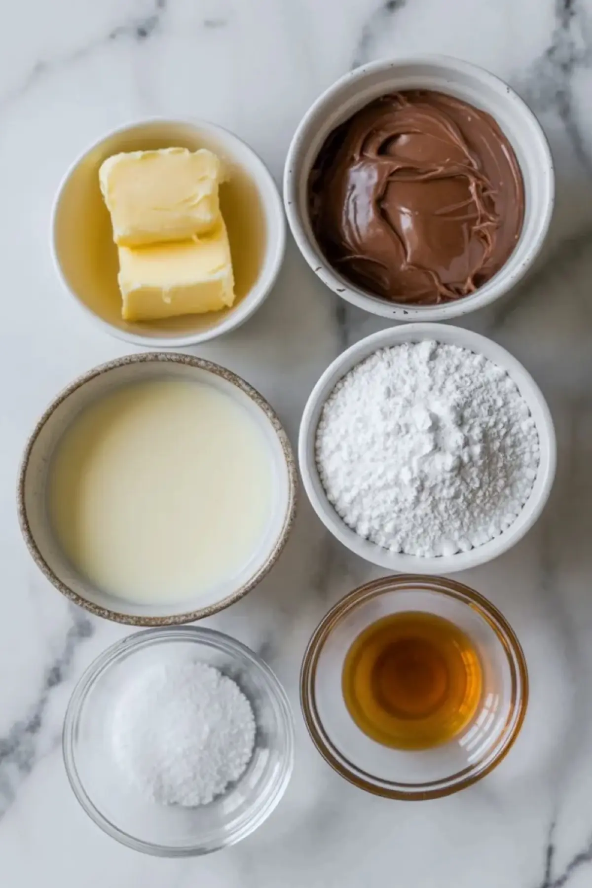 Ingredients for Nutella buttercream frosting arranged on white marble with butter, Nutella, powdered sugar, cream, vanilla, and salt in small bowls and spoons.