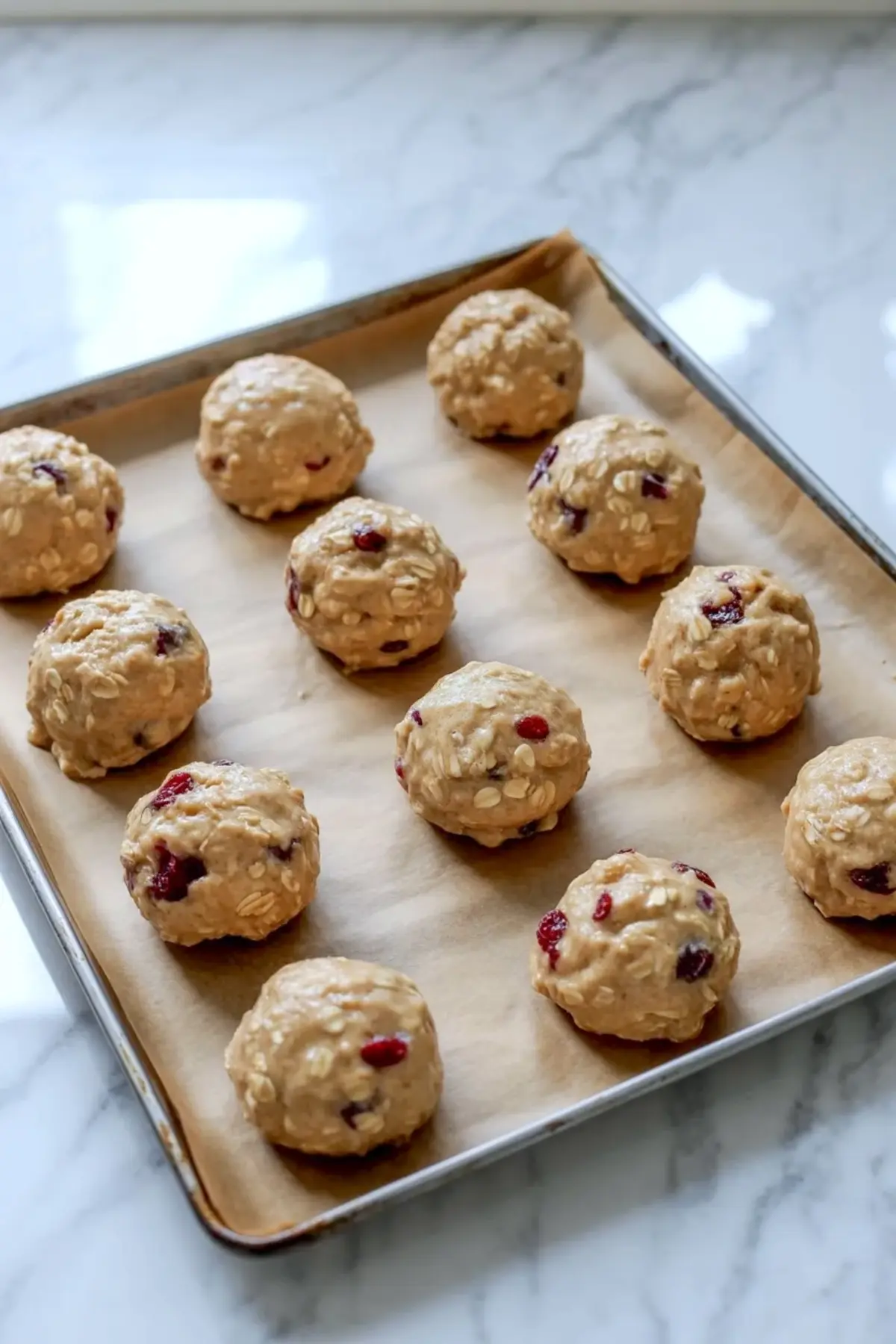 Scooped oatmeal cranberry cookie dough portions lined on a baking sheet before baking, with visible oats and dried cranberries throughout the dough.