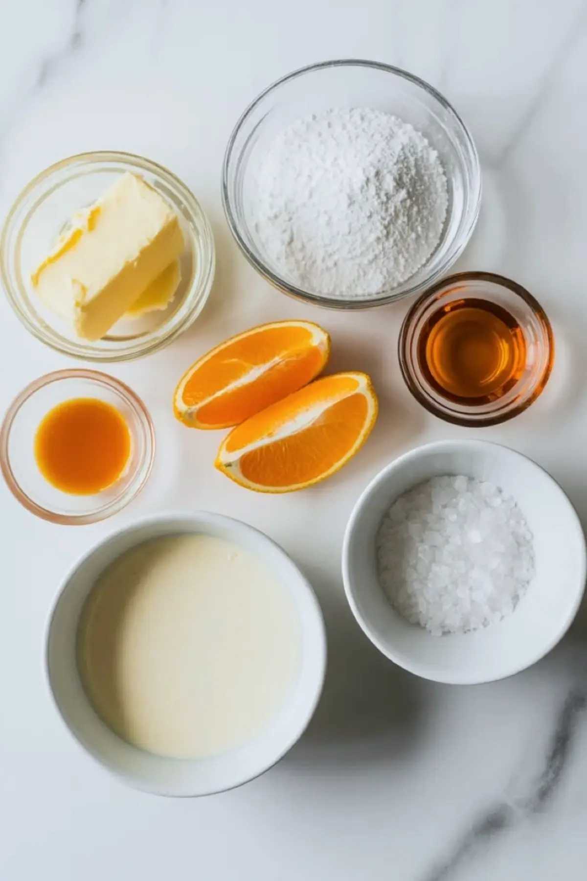 Ingredients for orange buttercream frosting are arranged on white marble with butter, powdered sugar, fresh orange, cream, vanilla, and salt.