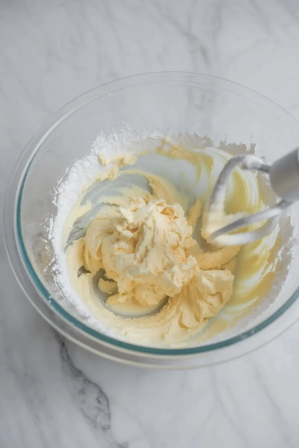Mixer beaters whip orange buttercream frosting in a glass bowl as the butter and sugar turn light and fluffy.