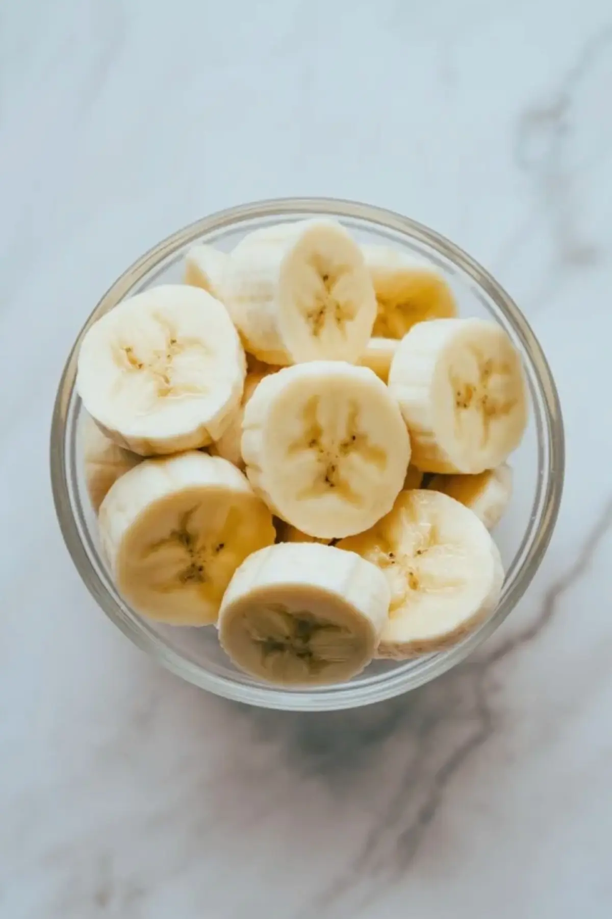 Sliced bananas for passion fruit banana juice packed into a clear glass jar on a light marble surface.