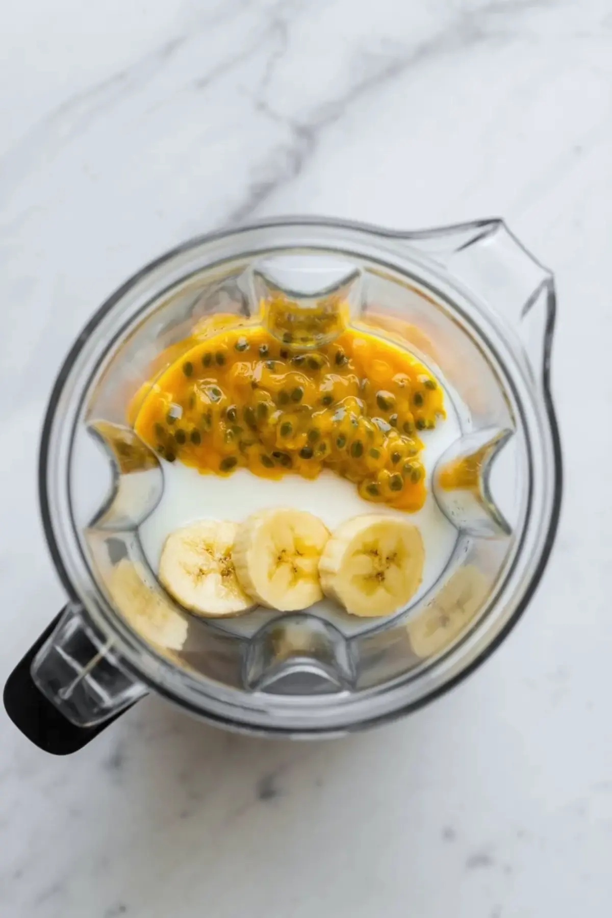 Blender jar filled with banana, passion fruit pulp, and liquid before blending into passion fruit banana juice on a white marble counter.