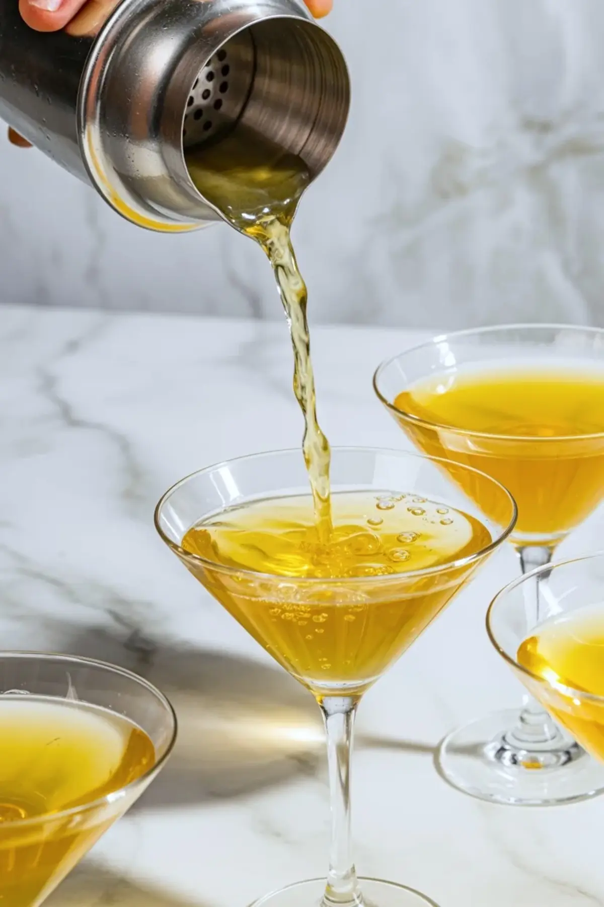 Passion fruit martini being poured from a metal shaker into a coupe glass, with other filled tropical cocktails nearby on white marble.