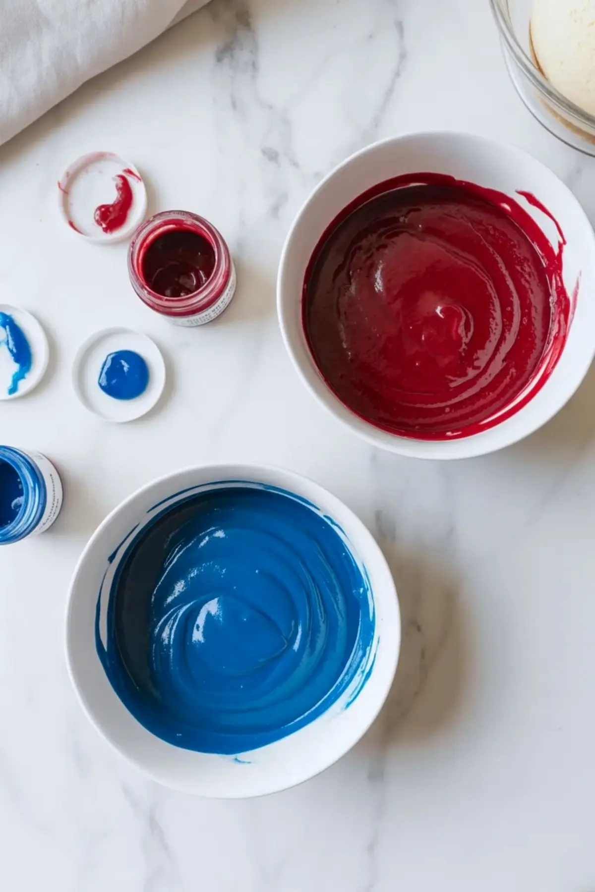 Red and blue batter for patriotic whoopie pies sits in separate bowls beside food coloring jars on a white marble surface.