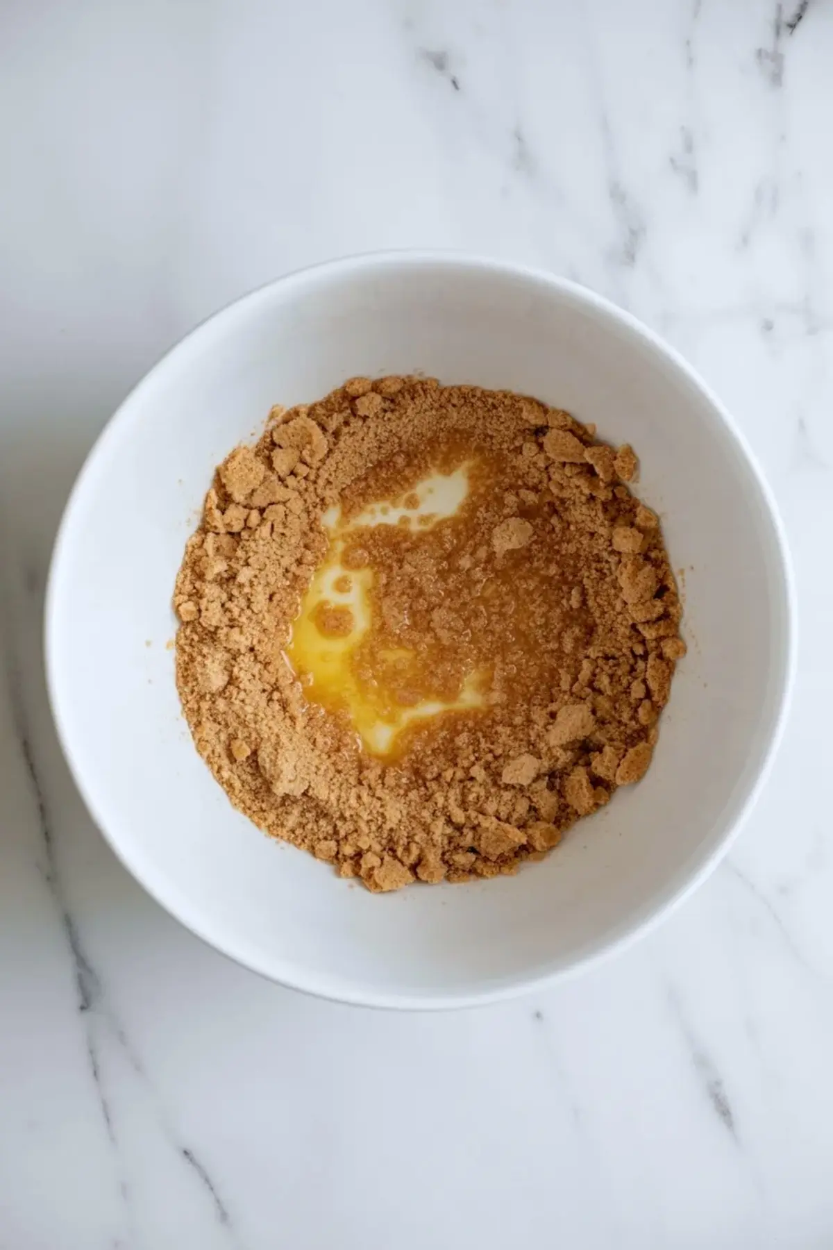 Graham cracker crumbs mixed with melted butter in a white bowl for pink lemonade pie crust on a marble surface.