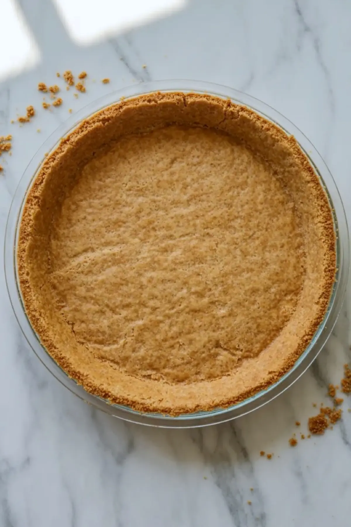 Prepared graham cracker crust for pink lemonade pie pressed into a clear pie dish on a white marble surface.