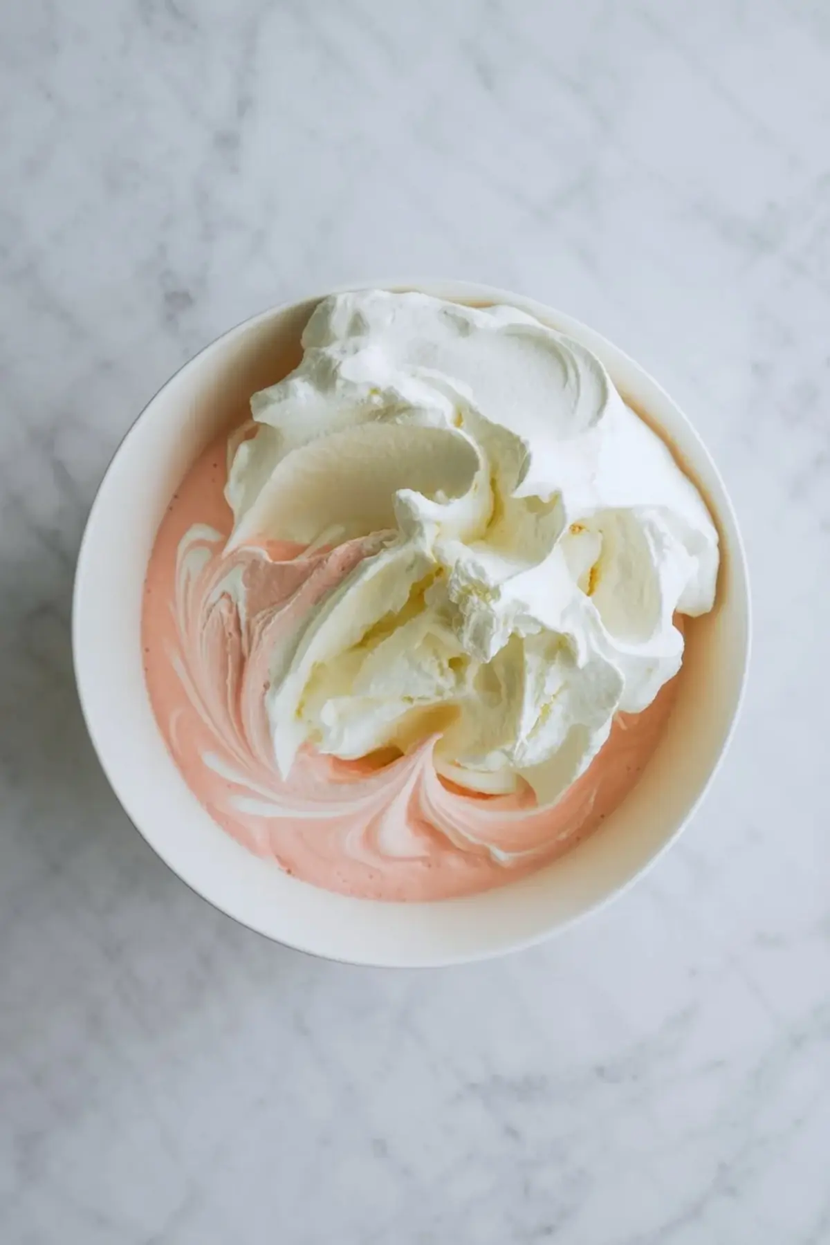 Pink lemonade pie filling in a white bowl with creamy pink mixture and whipped topping being folded together on a marble counter.