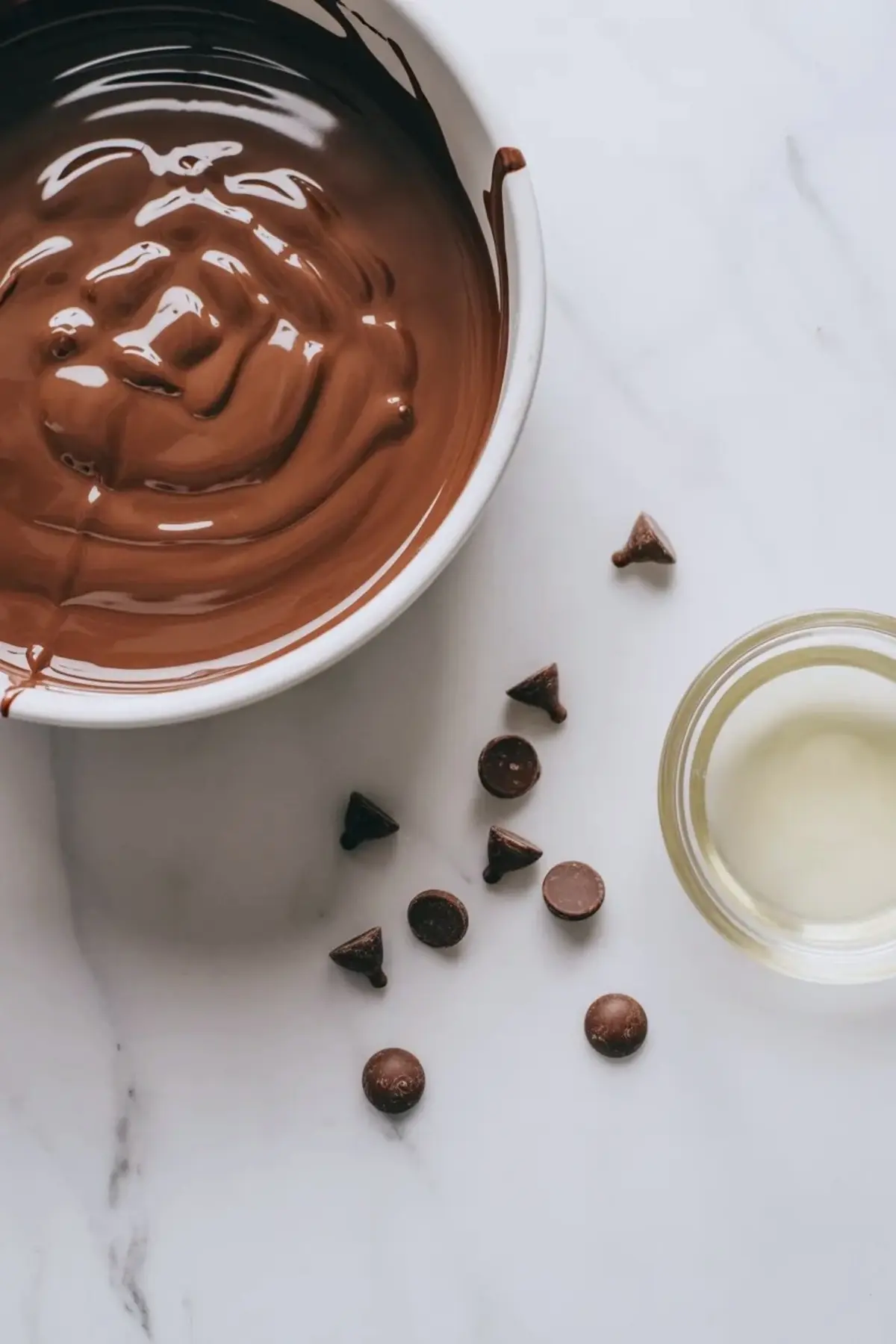 Melted chocolate topping for salted caramel cracker bites in a white bowl with chocolate chips and coconut oil beside it on a white marble surface.
