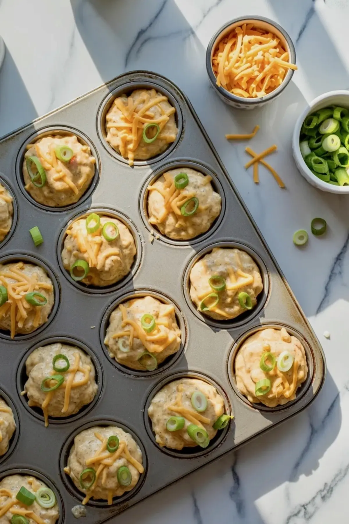 Sausage breakfast muffin batter portioned into a muffin pan and topped with cheddar cheese and green onions before baking.