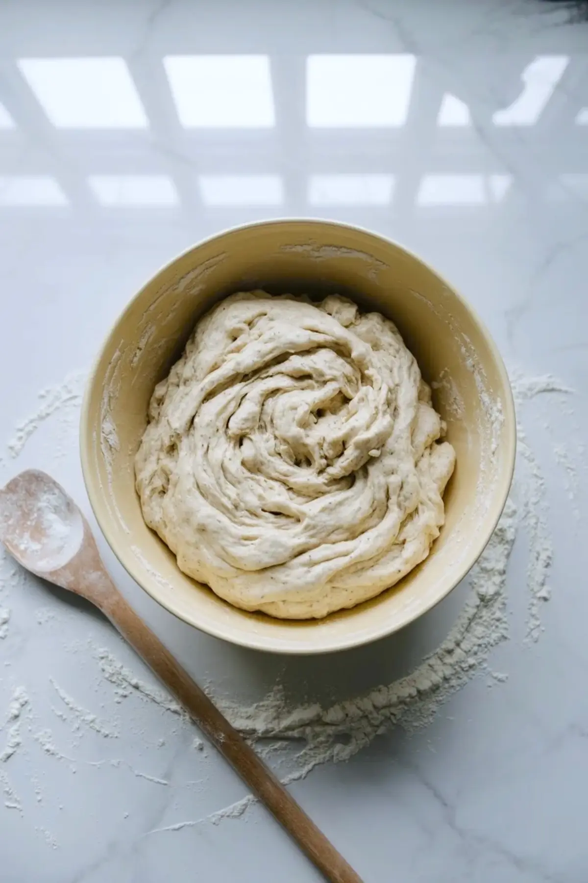 Freshly mixed sourdough bagel dough in a bowl with a wooden spoon on a floured marble work surface.