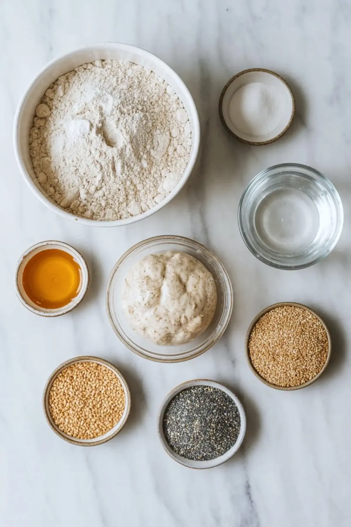 Ingredients for sourdough bagels arranged overhead with flour, starter, water, honey, salt, and topping seeds on a white marble surface.