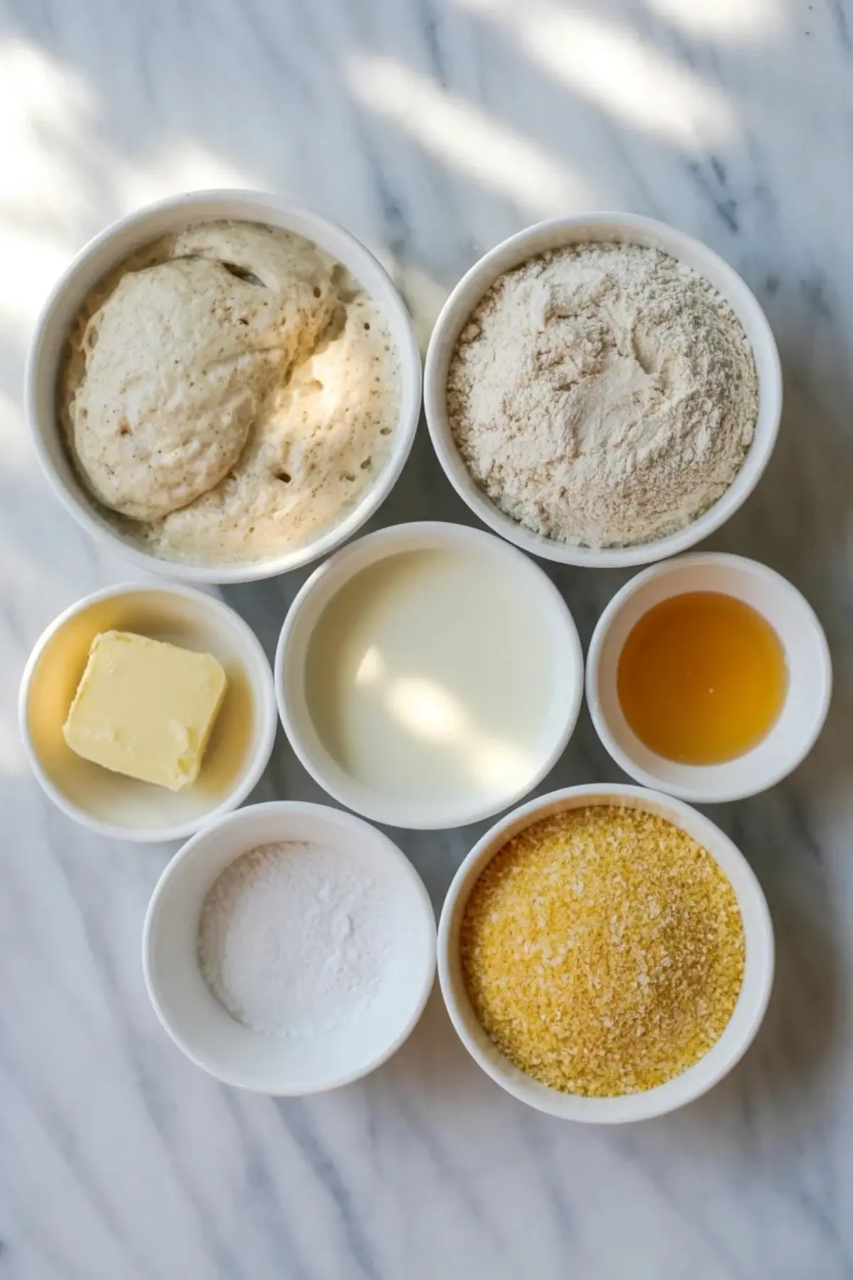 Ingredients for sourdough English muffins arranged overhead with sourdough starter, flour, milk, honey, butter, salt, and cornmeal.
