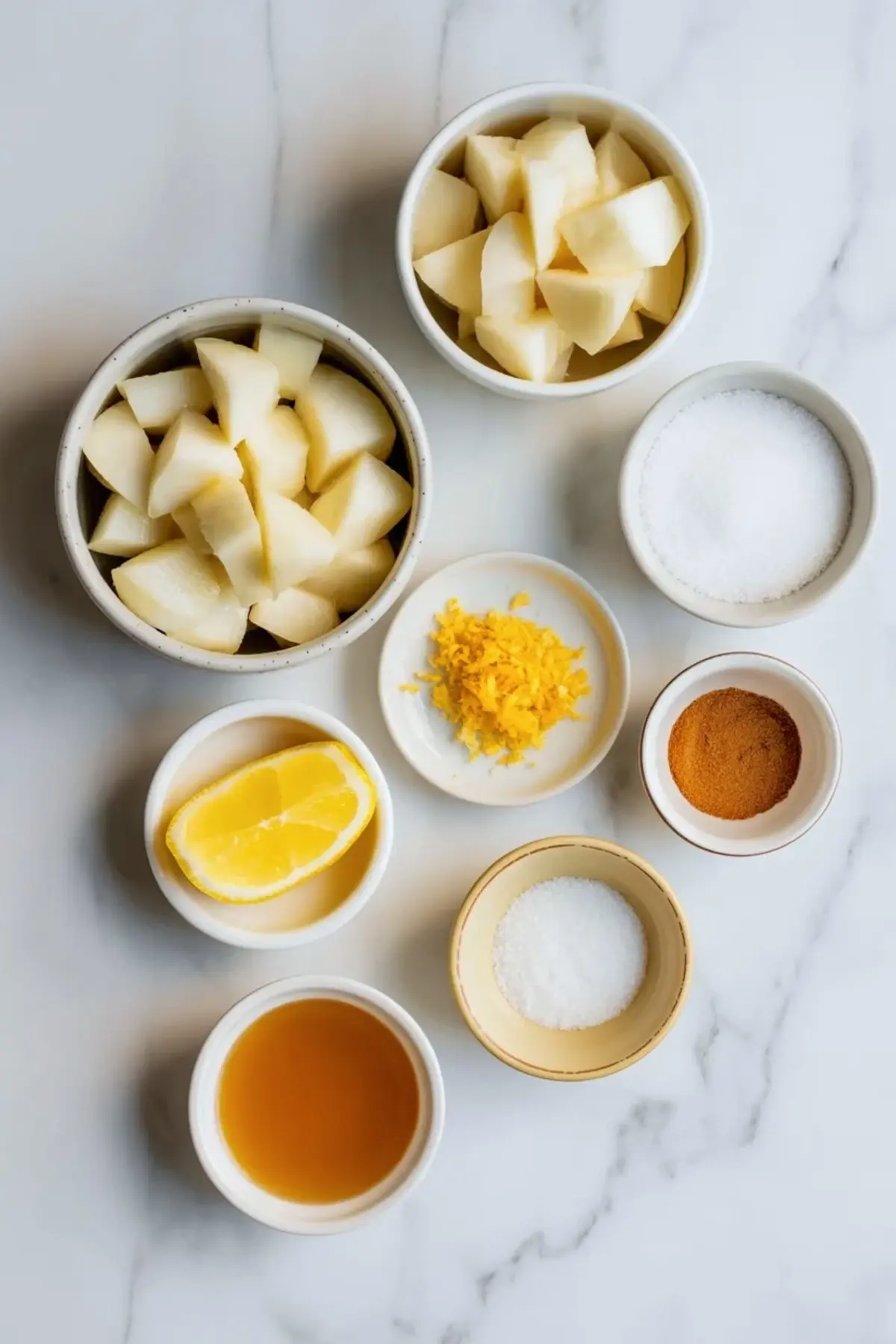 Ingredients for white peach jam recipe on white marble with chopped white peaches, sugar, lemon wedge, lemon zest, ground cardamom, salt, and a small bowl of liquid.