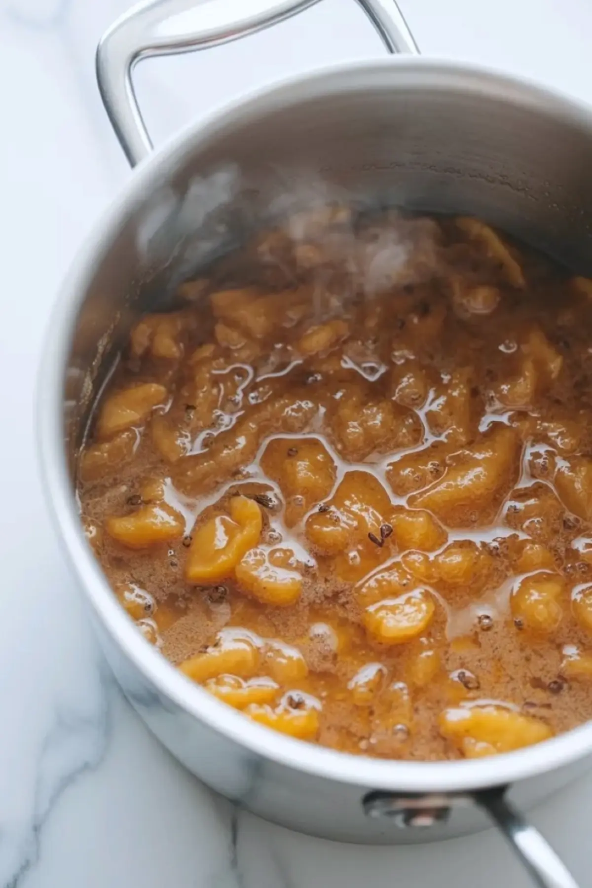 White peach cardamom jam cooking in a saucepan with bubbling peach mixture on a white marble counter.