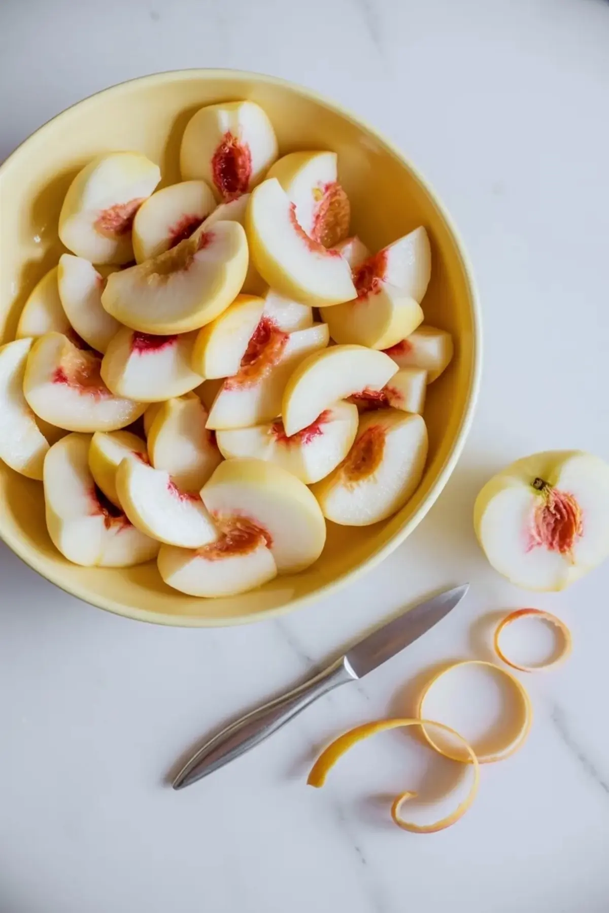 Prepared white peaches for jam in a yellow bowl with peeled peach slices, peach peels, and a small knife on a white marble work surface.
