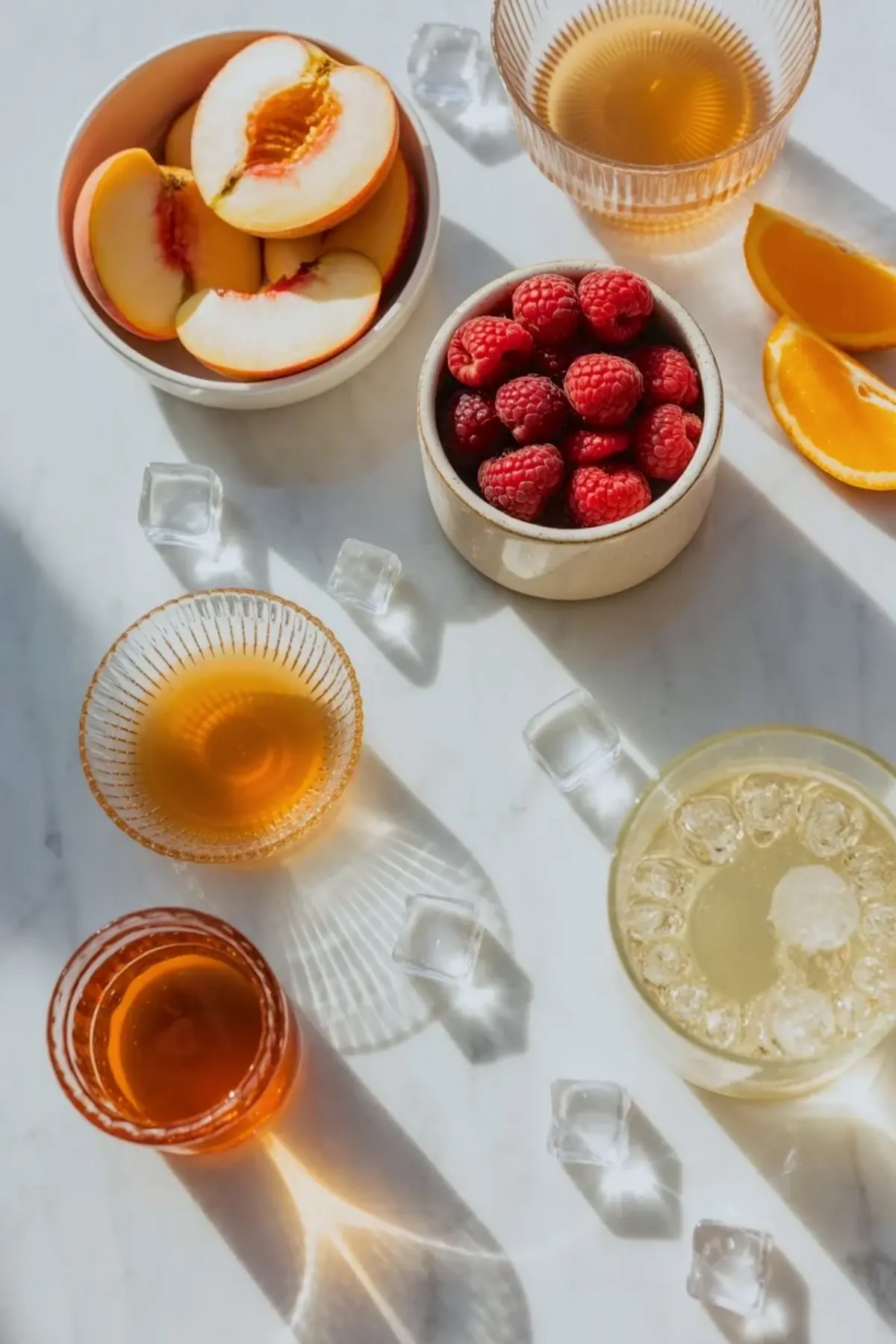 Ingredients for white peach sangria on white marble with sliced peaches, raspberries, orange wedges, ice, and glasses ready for serving.