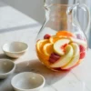 White peach sangria recipe shown in a clear glass pitcher with sliced peaches, orange, berries, and chilled white sangria on a white marble surface.