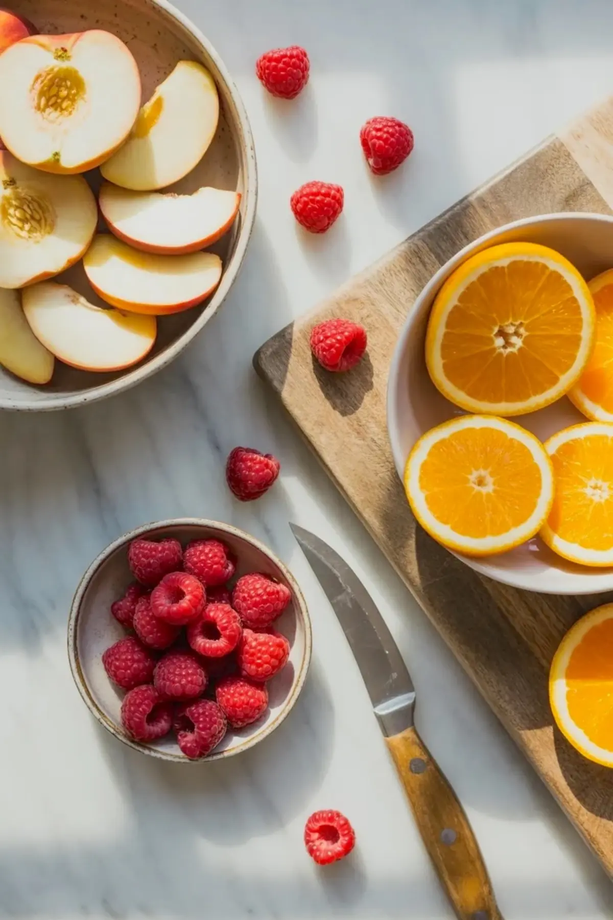 Fresh fruit for white peach sangria with sliced apples or peaches, orange slices, raspberries, and a knife on a white marble surface.