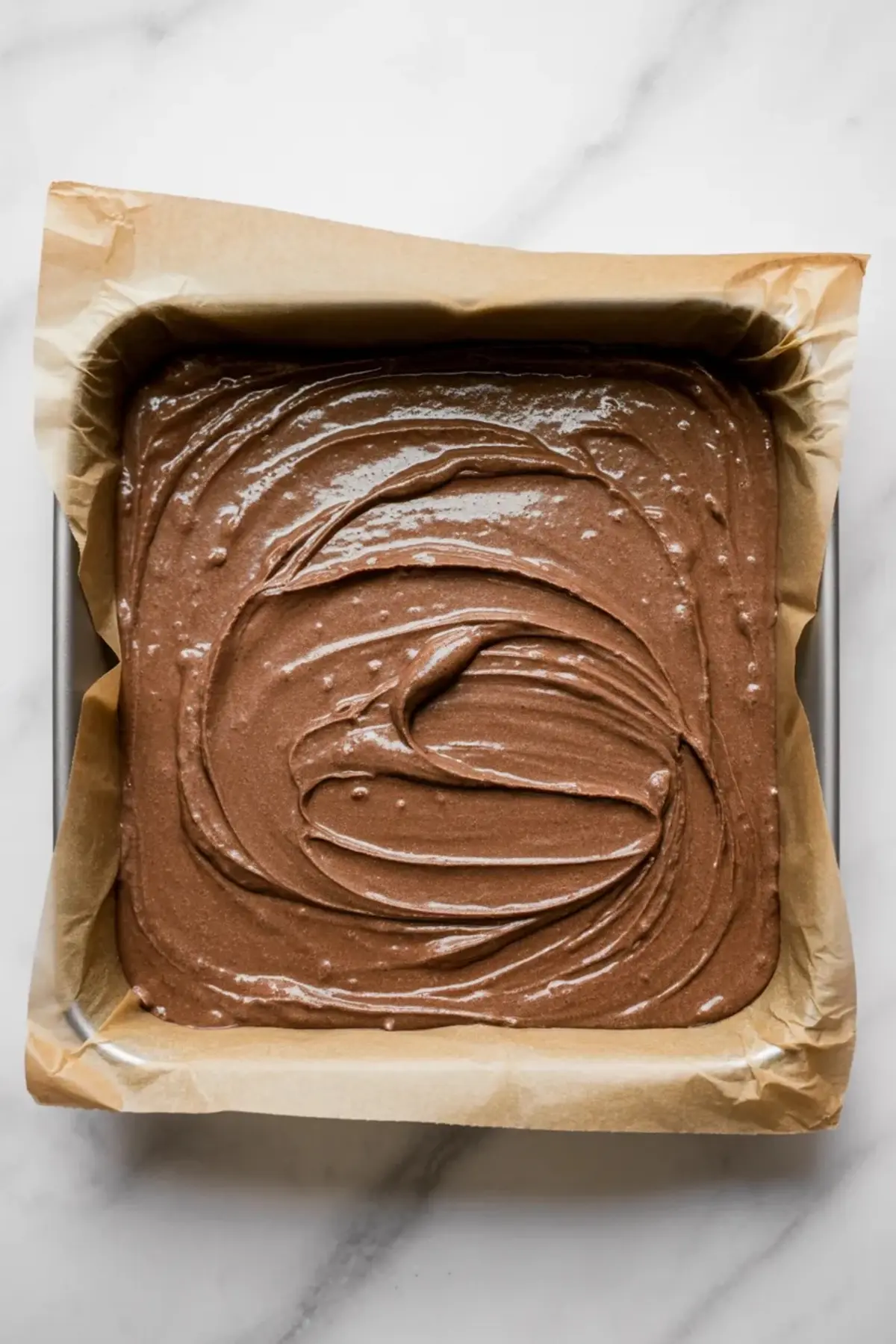 Chocolate brownie batter spread in a parchment lined square pan before baking the base for buckeye brownies.