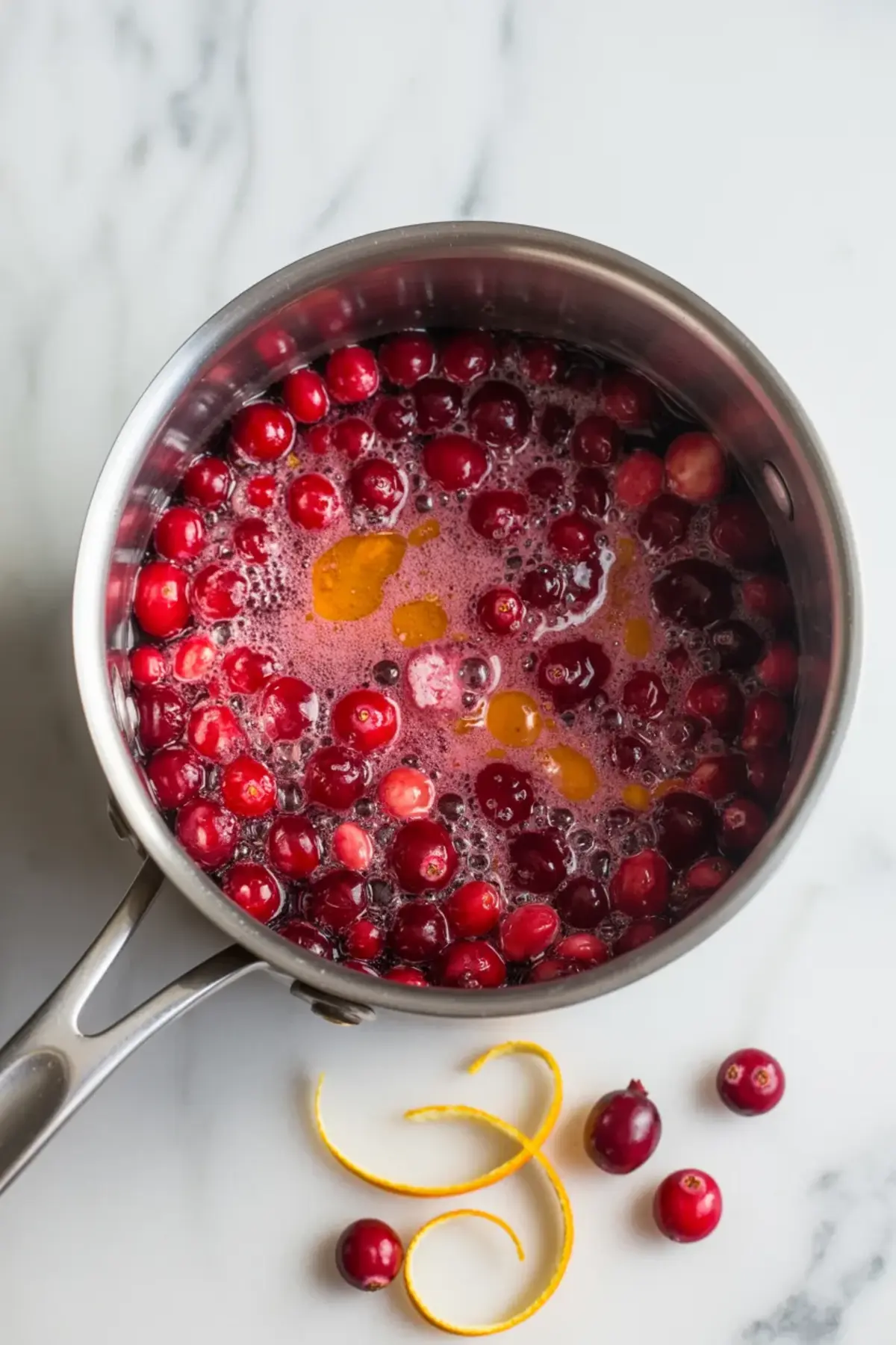 Fresh cranberries cooking in a saucepan for cranberry cream cheese dip with orange zest nearby on a white marble surface.