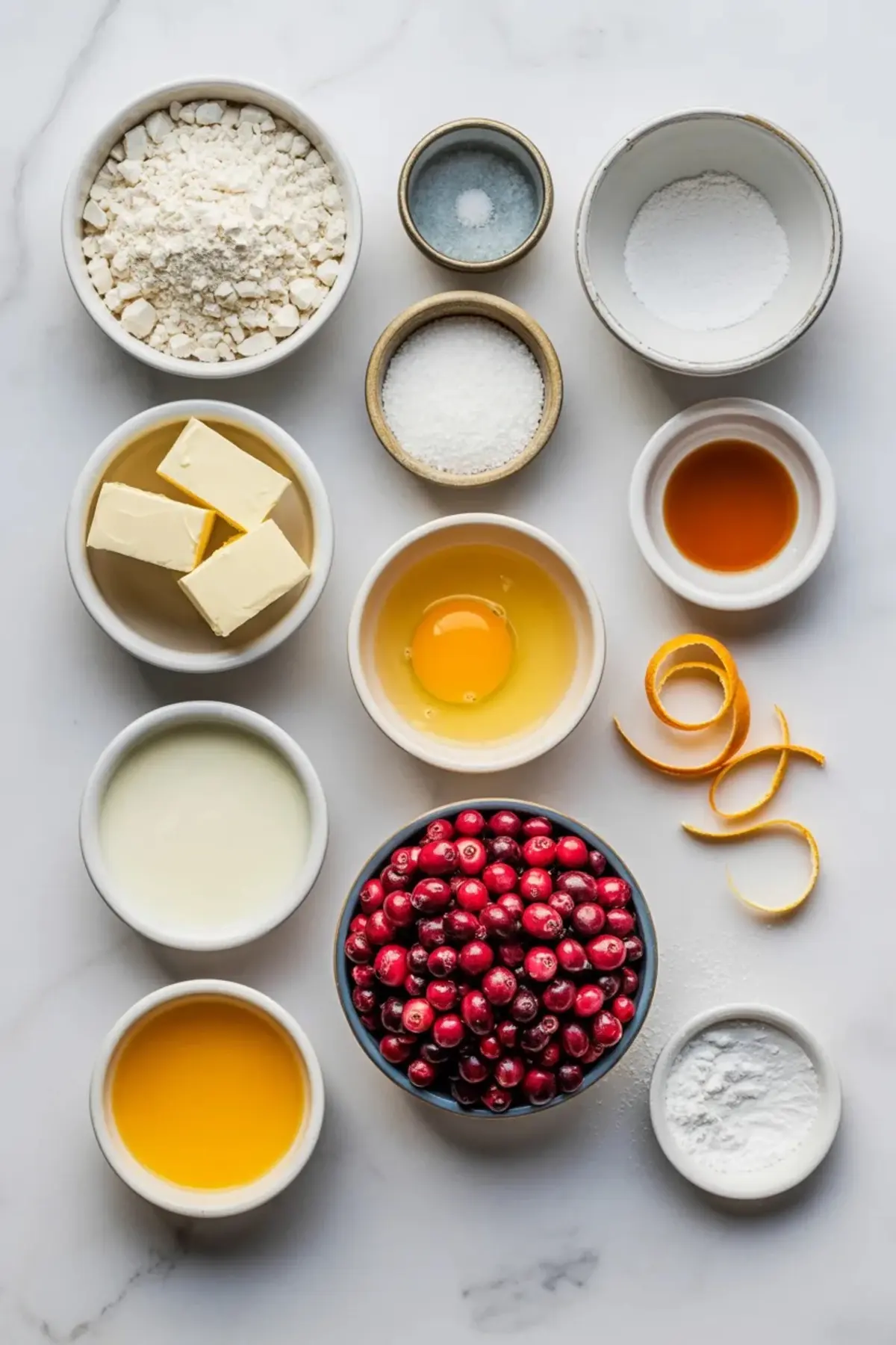 Measured cranberry orange scone ingredients arranged in bowls, including flour, sugar, butter, cranberries, egg, cream, orange juice, and orange zest.