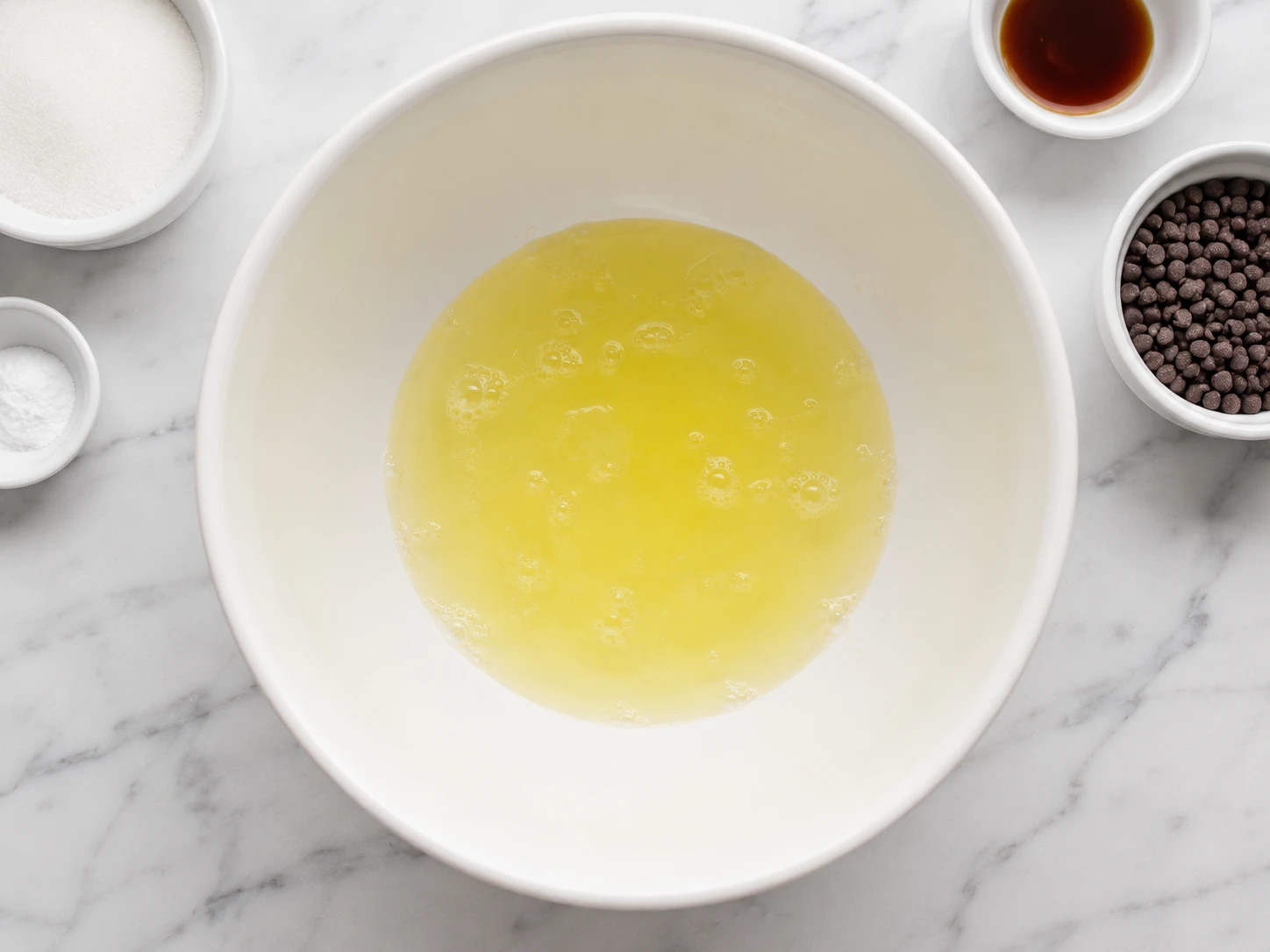 Raw egg whites in a large white mixing bowl, with small bowls of sugar, cream of tartar, vanilla and mini chocolate chips on a marble counter.