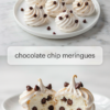 Chocolate chip meringues piped in tall swirls on a white plate, above two meringues broken open to show an airy white center full of mini chocolate chips.